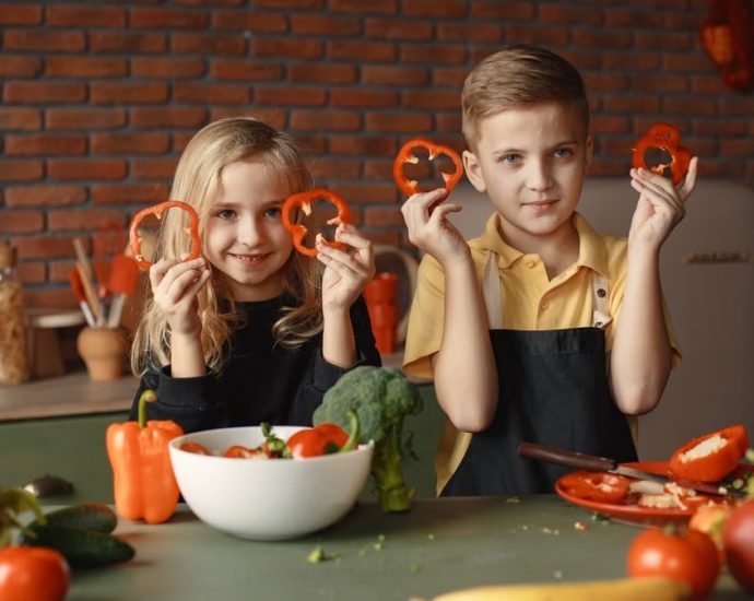 Two children holding capsicum slices while preparing vegetables in a cozy kitchen setting.