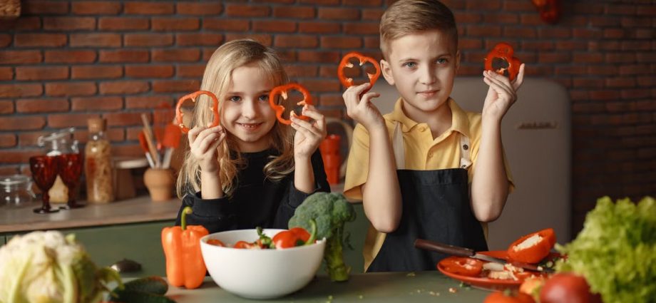 Two children holding capsicum slices while preparing vegetables in a cozy kitchen setting.
